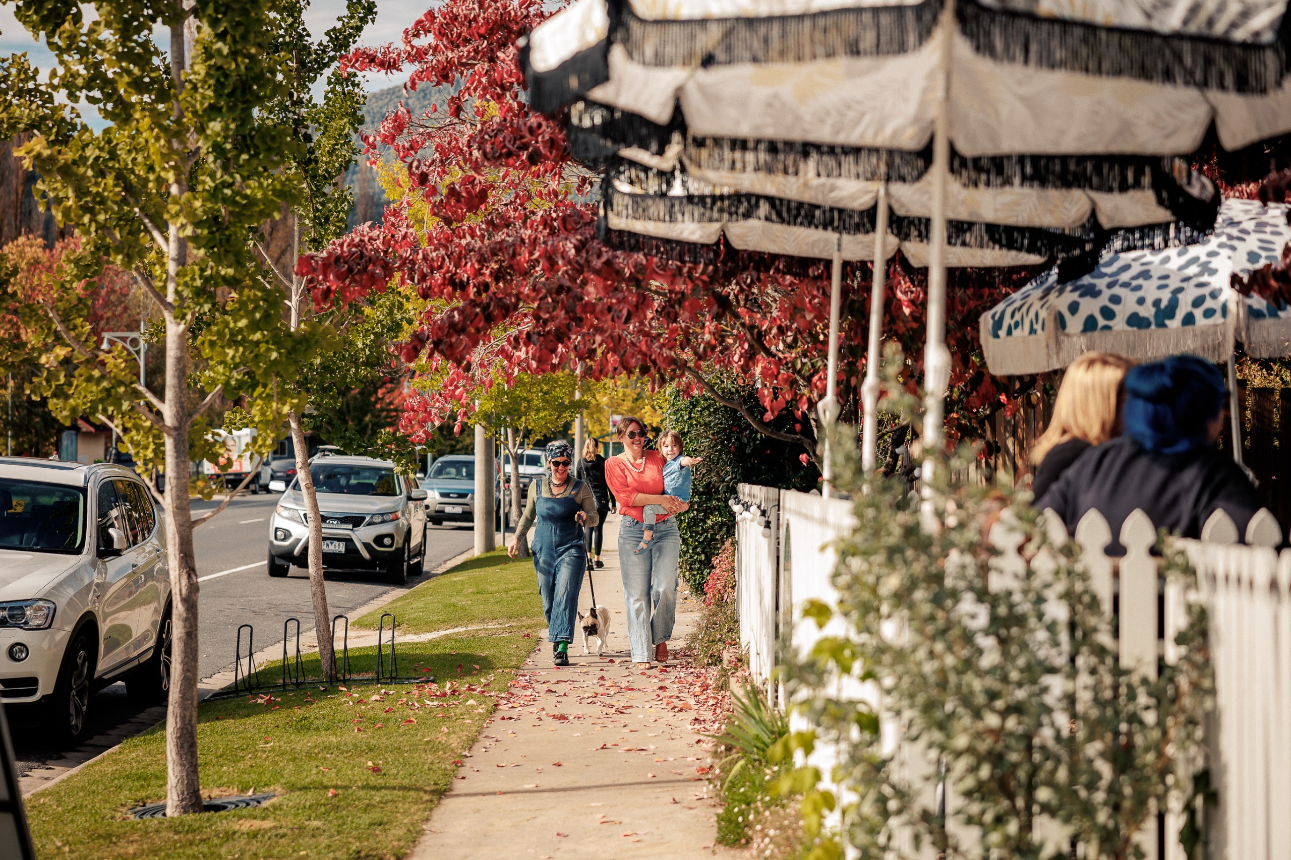 Two women walk along a footpath with shop fronts on the right hand side and a road on the left.