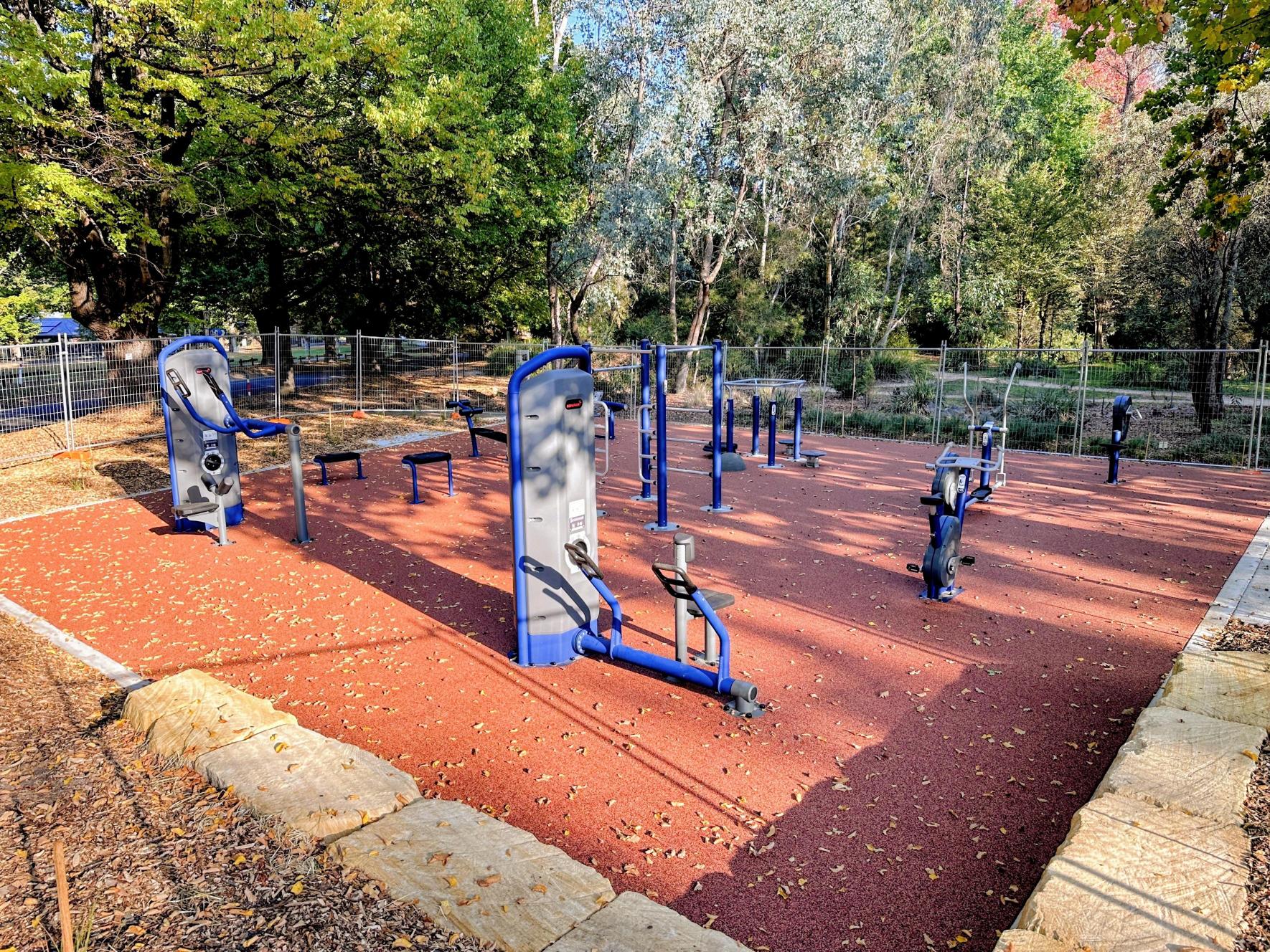 A variety of outdoor fitness equipment outside on top of an orange rubber surface, with green trees in the background.
