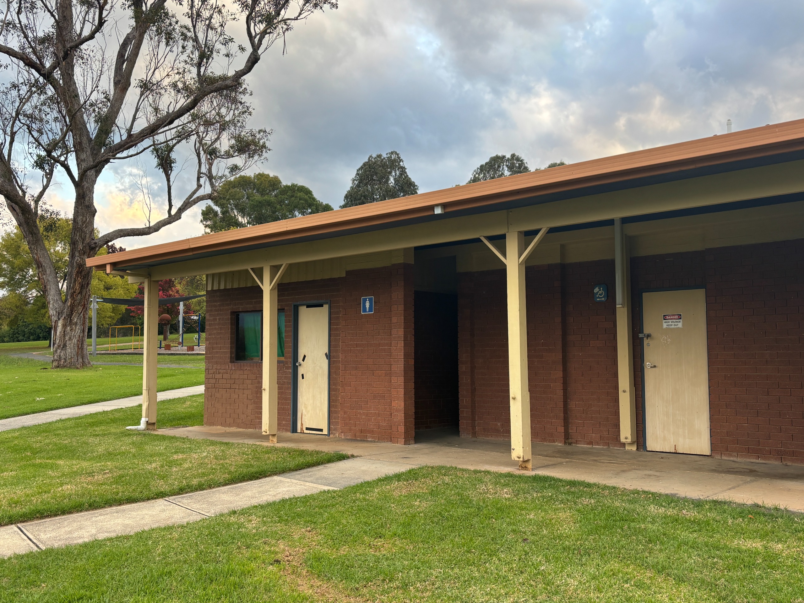 A red brick toilet building with cream doors and a lovely gumtree in the background.