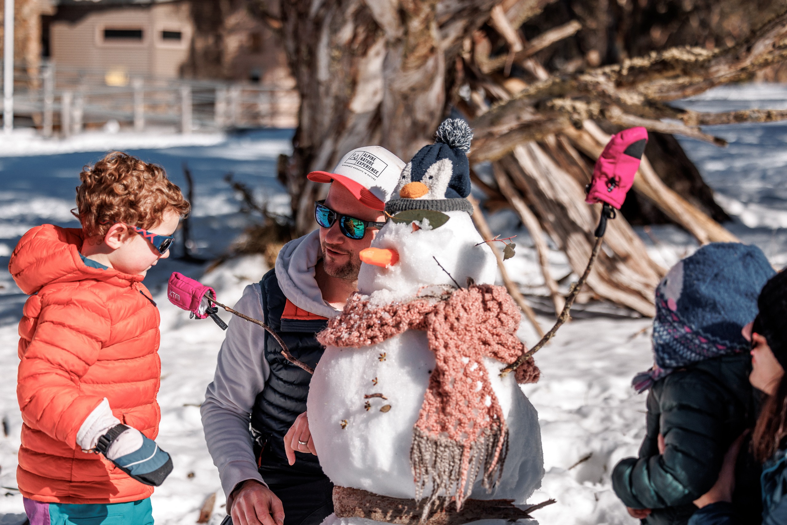 A father and his son build a snowperson. The snowperson is wearing a pink scarf and a penguin-shaped beanie.