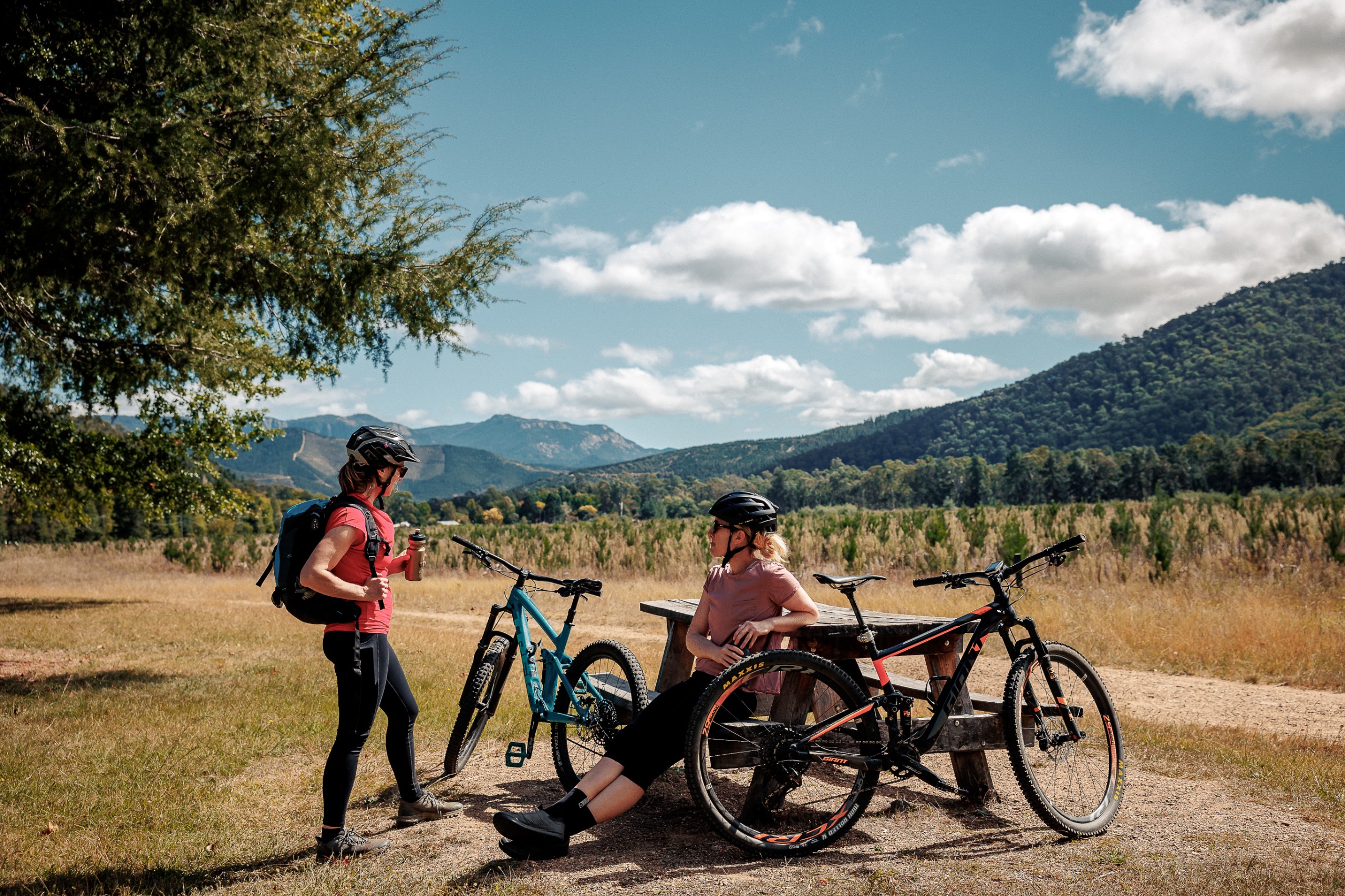 Two women are together with their pushbikes. One is standing, while the other is sitting at a picnic table. There are hills in the background.