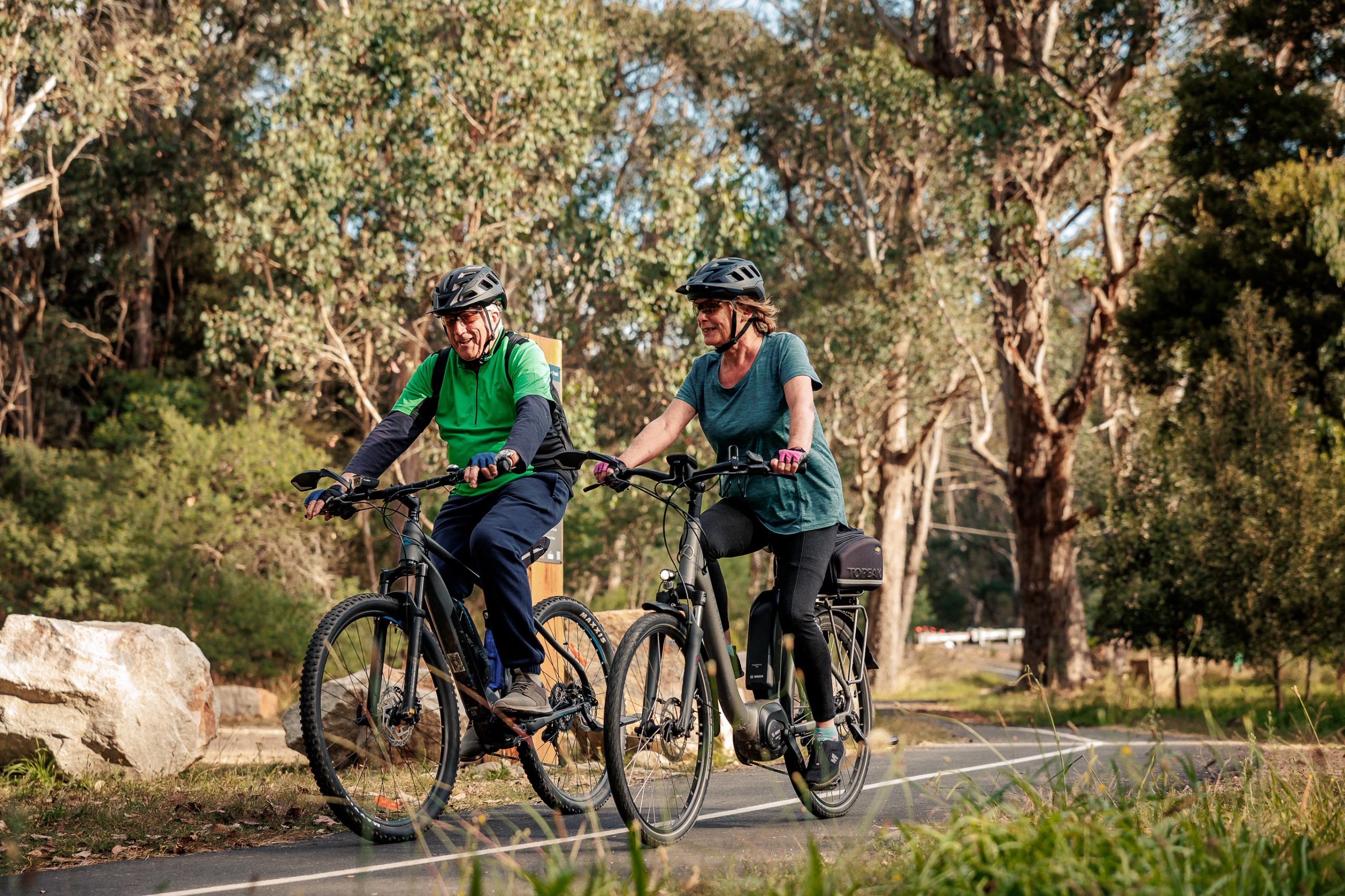 Two seniors ride their bikes along a path lined by mature trees.